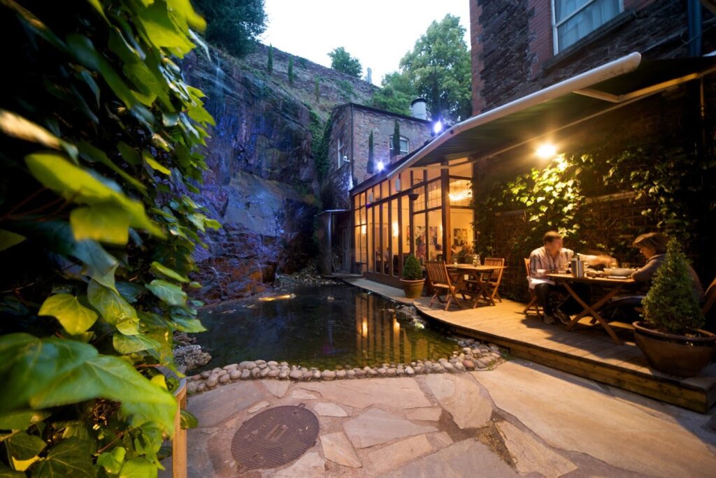 The open air terrace of Greene's Restaurant, with an artificial pond and a rocky wall lined with trees.