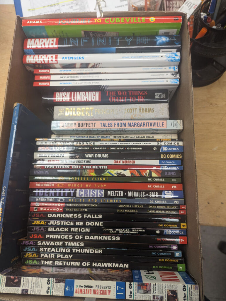 A Bankers box of books ready for the sale, containing graphic novels, Dilbert compilations, and a Rush Limbaugh book.