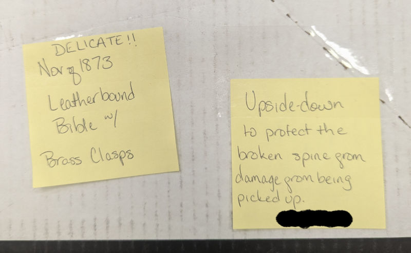 Two separate sticky notes. One reads, 'Delicate!! Nov of 1873, Leatherbound Bible w/ brass clasps' and the other reads, 'Upside-down to protect the broken spine from damage from being picked up.'