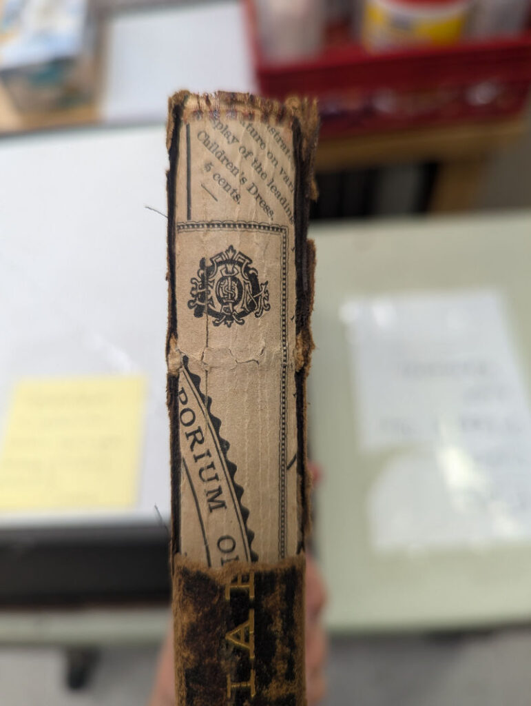 A closeup of the torn spine covering, revealing paper underneath, part of which advertises children's clothing for five cents.