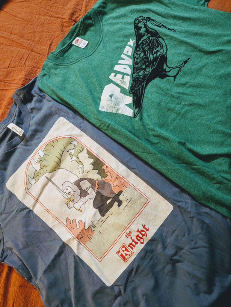 Two t-shirts: a blue one featuring the Noble Knight lying in wait for a dragon, and a green one with the Red Vox logo and a crow eating a worm.