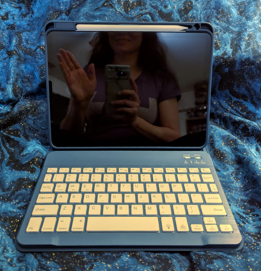 An iPad Air in a blue keyboard case; in the dark screen, the blogger is seen from the torso up, with her upper face hidden, waving with one hand while taking a selfie with a phone.