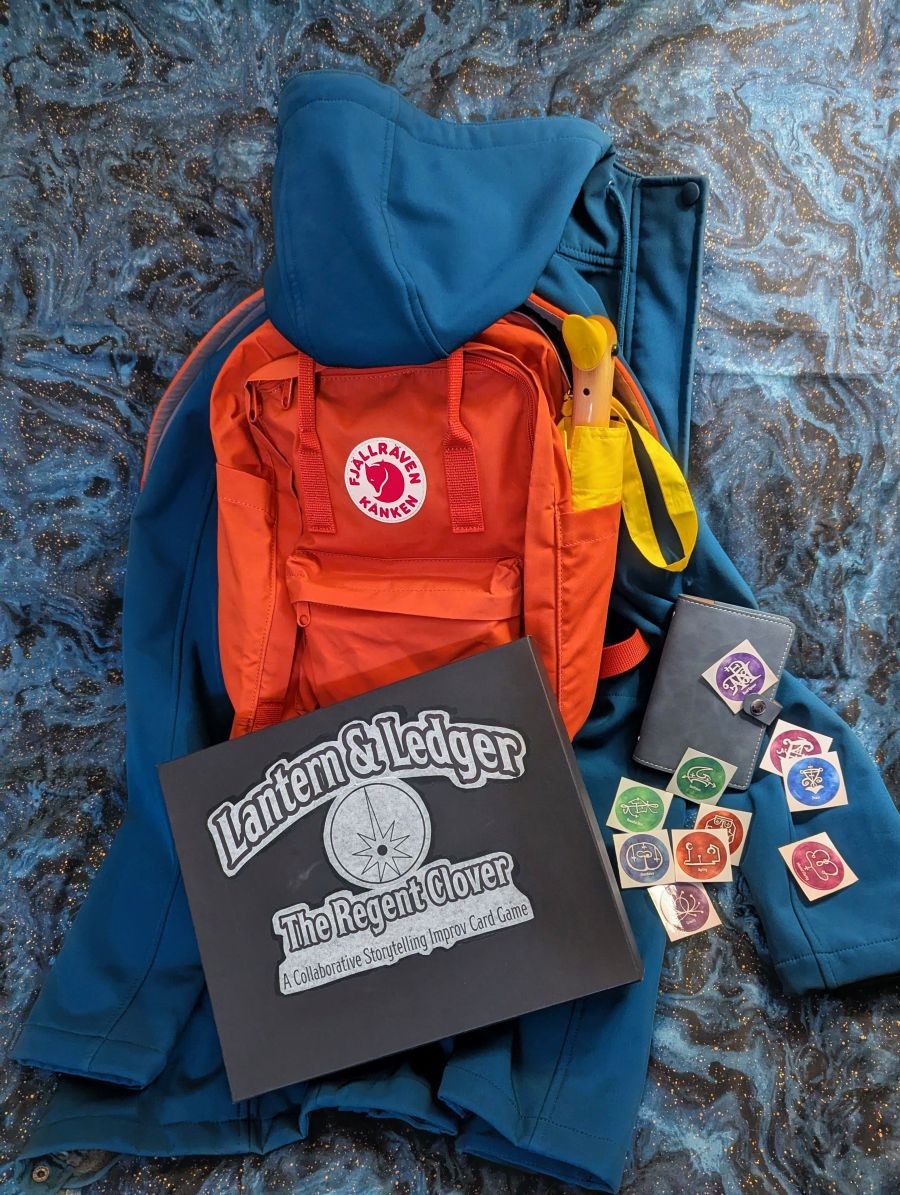A photographic setup featuring an orange Fjallraven backpack and a teal raincoat draped over it from behind. In the front is a black game box for Lantern & Ledger: The Regent Clover. To the right is a small blue binder with several round stickers of sigils spread around.