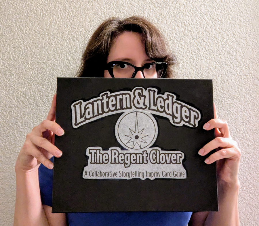 A photo of Ellie and the box for Lantern & Ledger: The Regent Clover. Ellie holds the box to conceal the lower part of her face, showing only her wavy brown hair, cat eye glasses, and eyes.